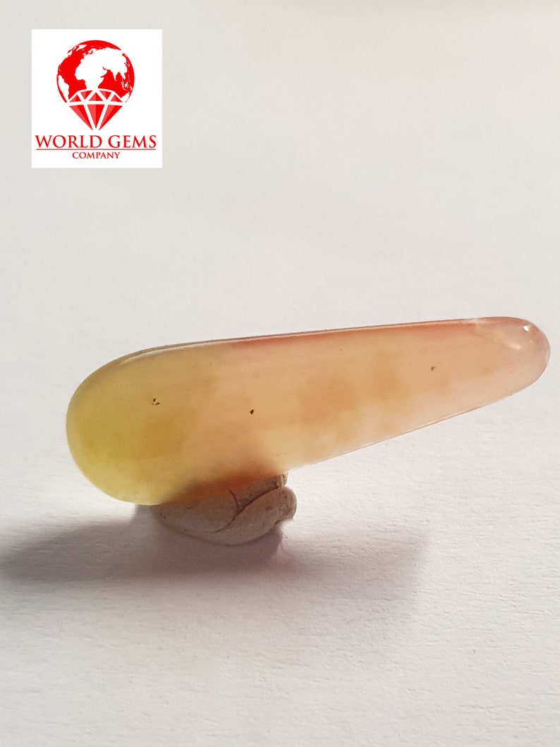May include: A long, smooth, light pink and yellow gemstone with a slightly pointed end. The gemstone is sitting on a small, grey, textured base.