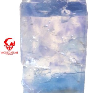 Peut inclure: Une pierre précieuse rectangulaire translucide de couleur bleu clair avec des fractures et des inclusions internes visibles. La pierre présente un dégradé de couleur allant d'un bleu pâle glacé au sommet à un bleu plus saturé à la base. Logo World Gems Company.