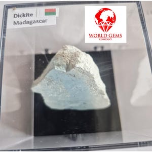 May include: A light-colored Dickite mineral specimen from Madagascar, displayed in a clear case. The mineral has a rough, textured surface with hints of blue. The case includes a label with the mineral's name and origin.