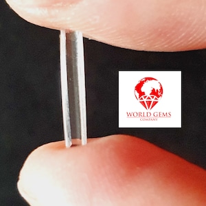 Op de afbeelding: Een heldere, rechthoekige, gefacetteerde edelsteen die tussen twee vingers wordt vastgehouden. De edelsteen is ongeveer 2,5 cm lang. De afbeelding is gelabeld met "WORLD GEMS COMPANY".