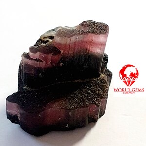 Könnte beinhalten: Ein roher, natürlicher, rosa und schwarzer Turmalinkristall mit Streifen. Der Kristall ist etwa 5 cm lang und 2,5 cm breit.