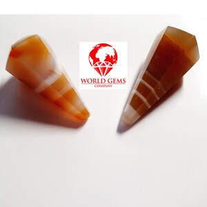 Peut inclure: Deux pendentifs en pierres précieuses en agate orange et blanche polie. Les pierres précieuses sont en forme de cône avec un dessus facetté et un motif rayé. Le logo "WORLD GEMS COMPANY" est visible en arrière-plan.