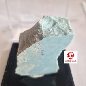 May include: A rough-cut, light blue-green mineral specimen, possibly a gemstone, displayed on a black base. The stone has a textured surface and a lighter, more weathered section. The World Gems Company logo is visible.