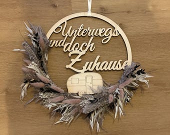 Könnte beinhalten: Ein hölzerner, kreisförmiger Kranz mit dem deutschen Satz "Unterwegs und doch Zuhause" (Unterwegs und doch zu Hause). Der Kranz zeigt eine Holzillustration eines Wohnwagens und ist mit getrockneten Blumen in Rosa-, Grau- und Weißtönen verziert.