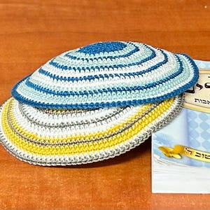 Può includere: Due kippah all'uncinetto, una a strisce blu e bianche e l'altra a strisce gialle, bianche e grigie. Le kippah sono appoggiate su un libro con testo ebraico.