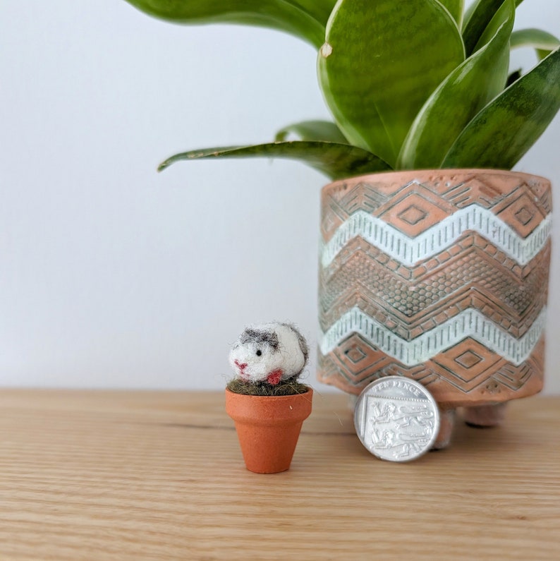 A small, white and grey felt guinea pig figurine sitting in a terracotta pot. The pot is on a wooden surface next to a potted plant.