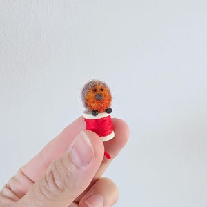 May include: A small, orange and grey felt animal sitting on a spool of red thread. The animal has a small black nose and eyes.