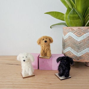 May include: Three miniature felted dog figurines in white, tan, and black, each on a small wooden base. The tan dog is perched on a pink block. A potted plant with green leaves is in the background.