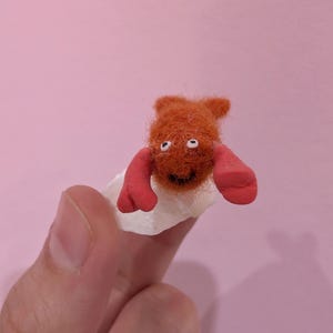 May include: A handmade orange felt creature with red arms and white eyes. The creature is attached to a white object and held against a pink background. The creature has a smiling mouth.