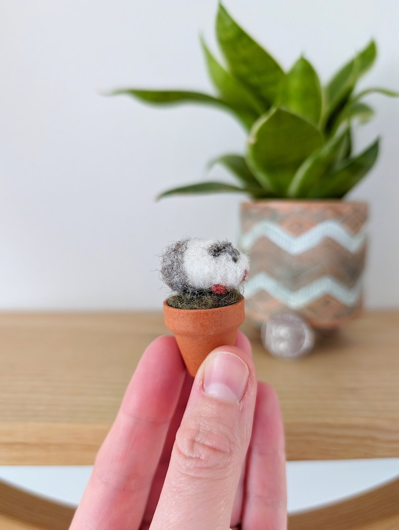 May include: A small, hand-felted animal figurine in a miniature terracotta pot. The animal is white with black and gray markings and red accents. The pot is filled with green felt.