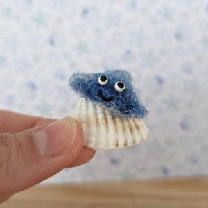 May include: A blue felt seashell with a smiling face and black eyes. The seashell is sitting on a white seashell.