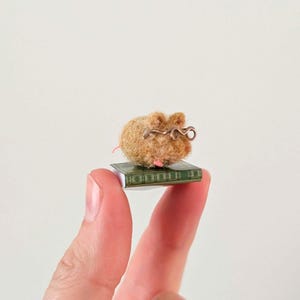 A persons fingers holding a Needle felt mouse wearing spectacles sat on top of a book. Miniature mouse gift suitable for book lovers.