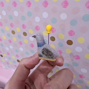 May include: A handmade felt pigeon figurine with gray, blue, and brown details, holding a yellow balloon. The bird is mounted on a small wooden base. The background features a pink polka dot pattern.