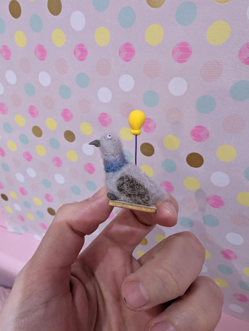 May include: A handmade felt pigeon figurine with gray, blue, and brown details, holding a yellow balloon. The bird is mounted on a small wooden base. The background features a pink polka dot pattern.
