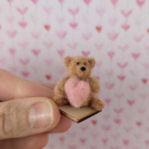 May include: A miniature brown teddy bear with a pink heart on its chest, sitting on a small wooden base. The background features a pattern of pink hearts. The bear is held by a person.
