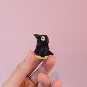 May include: A small, black bird ornament with a white eye and a black beak. The bird sits on a small, round, beige base. The figurine is made of a fuzzy material.