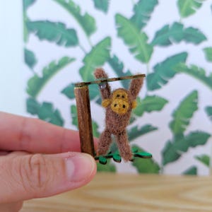 May include: A brown felt monkey figurine hanging from a wooden branch with green leaves. The monkey has a yellow face and is holding onto the branch with its hands.
