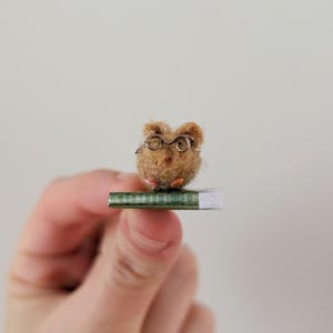 A persons fingers holding a Needle felt mouse wearing spectacles sat on top of a book. Miniature mouse gift suitable for book lovers.