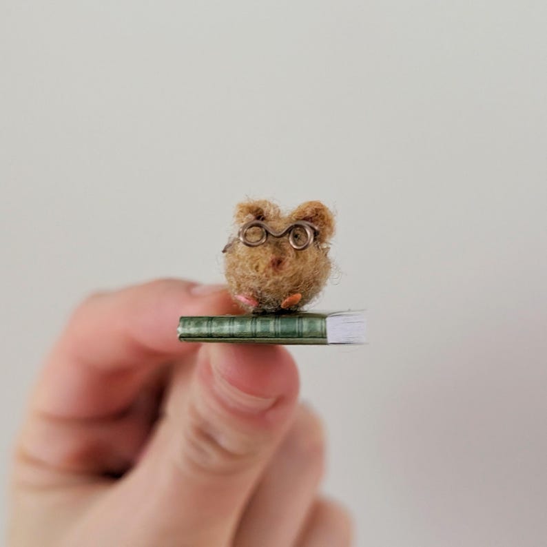 A persons fingers holding a Needle felt mouse wearing spectacles sat on top of a book. Miniature mouse gift suitable for book lovers.