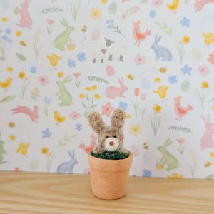 May include: A brown bunny rabbit with a white nose and green eyes is peeking out of a terracotta pot. The pot is filled with green moss. The bunny is sitting on a wooden surface with a white background featuring a repeating pattern of pastel coloured bunnies, chicks, flowers, and eggs.