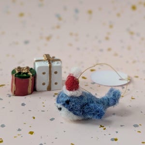 May include: A needle-felted blue whale ornament wearing a small red and white Santa hat, accompanied by two miniature gift boxes, one red with a green lid and gold ribbon, and one white with gold dots and a gold ribbon, featuring a gold hanging string.