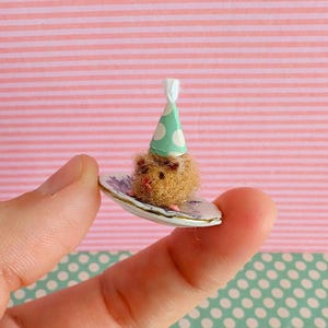May include: A tiny, brown, fluffy creature wearing a green party hat with white spots, resting on a miniature plate with a floral pattern. The plate is held between fingers, highlighting the small size of the whimsical object.