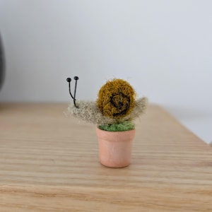 May include: A handcrafted felt snail figure with a brown shell and a light green body, sitting in a small terracotta pot filled with green felt. The snail has black antennae. The figure is on a wooden surface.