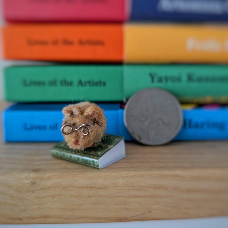 Needle felt mouse wearing spectacles sat on top of a book. With a background of books. Miniature mouse gift.