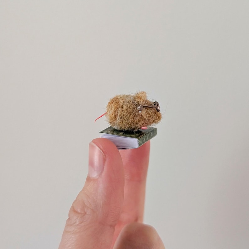 A persons fingers holding a Needle felt mouse wearing spectacles sat on top of a book. Miniature mouse gift suitable for book lovers.