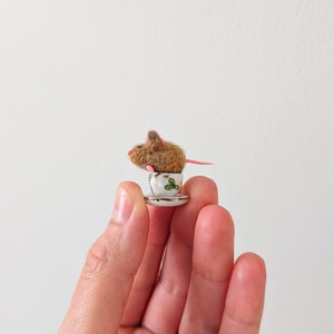 May include: A small brown mouse figurine sitting in a white teacup with green floral designs. The teacup is on a white saucer.