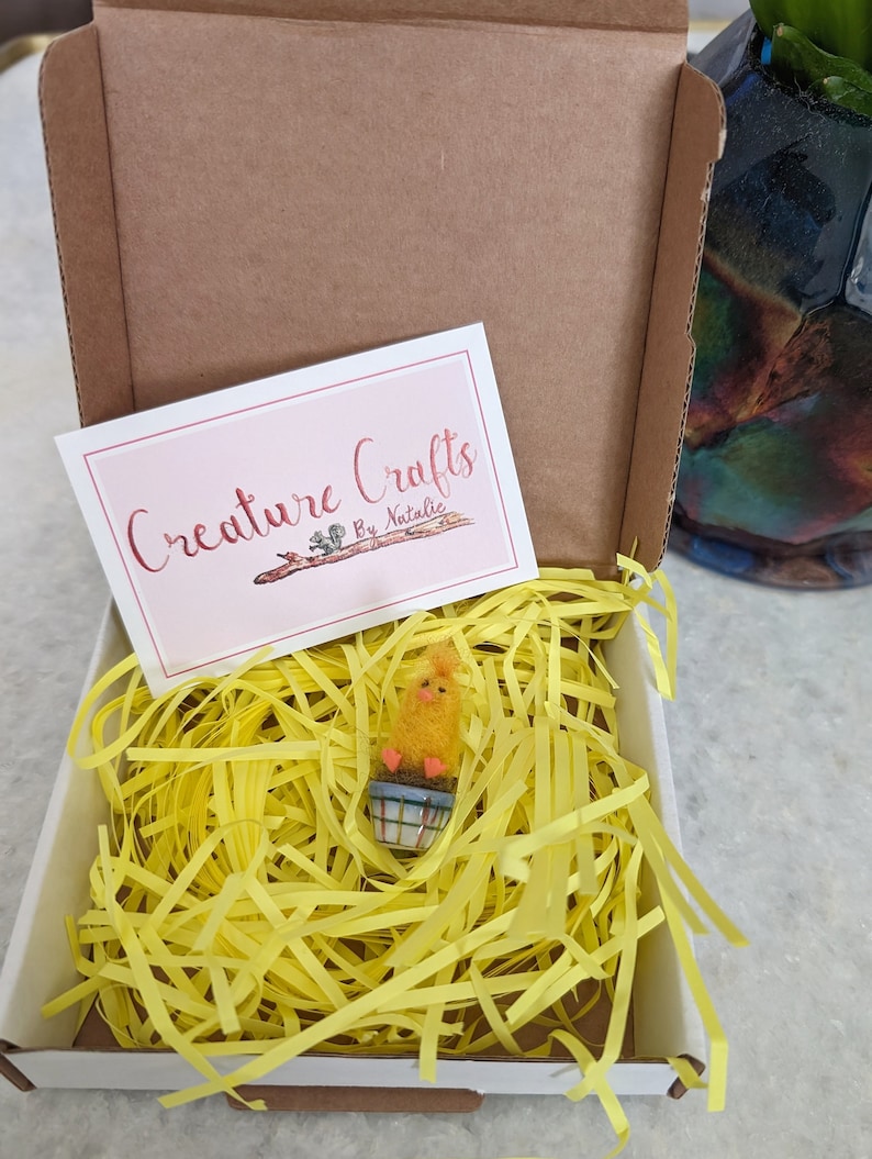 Puede incluir: Un peque&ntilde;o pollito amarillo en una maceta se encuentra en una caja blanca llena de papel amarillo triturado. La caja tiene una tarjeta en la parte superior que dice "Creature Crafts by Natalie".