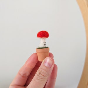 Puede incluir: Un peque&ntilde;o hongo de fieltro rojo y blanco en una maceta de terracota. El hongo tiene una cara sonriente.