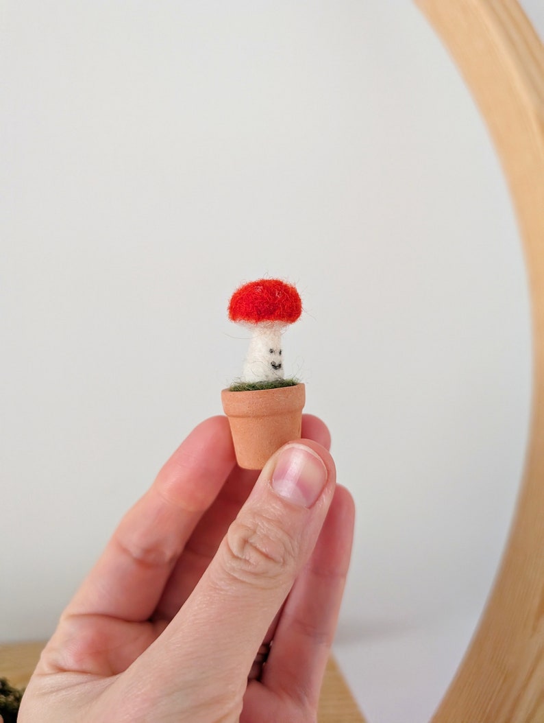 Puede incluir: Un peque&ntilde;o hongo de fieltro rojo y blanco en una maceta de terracota. El hongo tiene una cara sonriente.