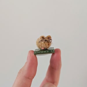 A persons fingers holding a Needle felt mouse wearing spectacles sat on top of a book. Miniature mouse gift suitable for book lovers.