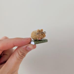 A persons fingers holding a Needle felt mouse wearing spectacles sat on top of a book. Miniature mouse gift suitable for book lovers.