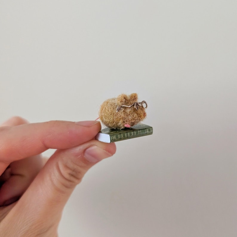 A persons fingers holding a Needle felt mouse wearing spectacles sat on top of a book. Miniature mouse gift suitable for book lovers.