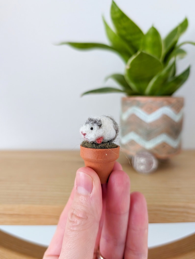A small, white and grey, needle felted guinea pig figurine sitting in a terracotta pot. The pot is small and has green wool around the rim.