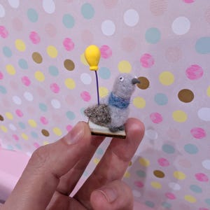 May include: A miniature felt pigeon figurine with a blue scarf and a yellow balloon. The bird is gray with a brown back and a small wooden base. The background is a pink surface with colorful polka dots.
