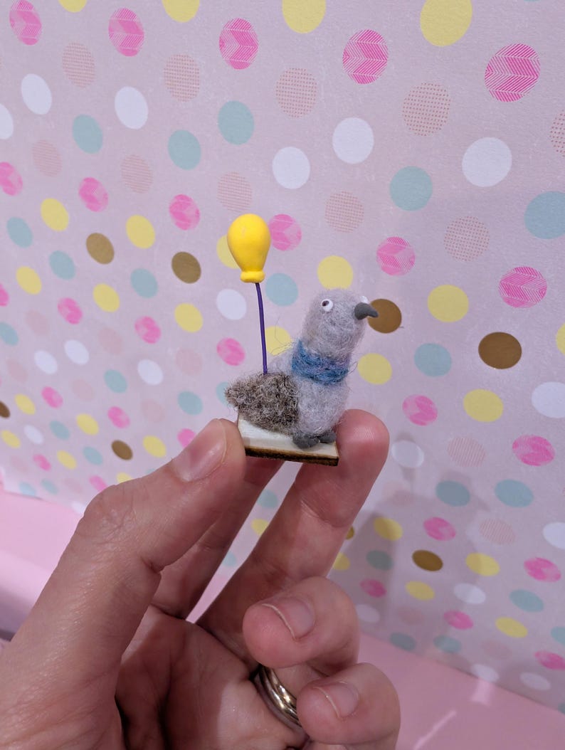 May include: A miniature felt pigeon figurine with a blue scarf and a yellow balloon. The bird is gray with a brown back and a small wooden base. The background is a pink surface with colorful polka dots.