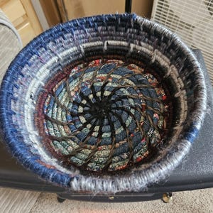 Op de afbeelding: Handgemaakte ronde mand met een spiraalvormig ontwerp. De mand is gemaakt van denim en andere stoffen in tinten blauw, grijs en bordeaux. De binnenkant heeft een spiraalpatroon met donkerbruin touw.