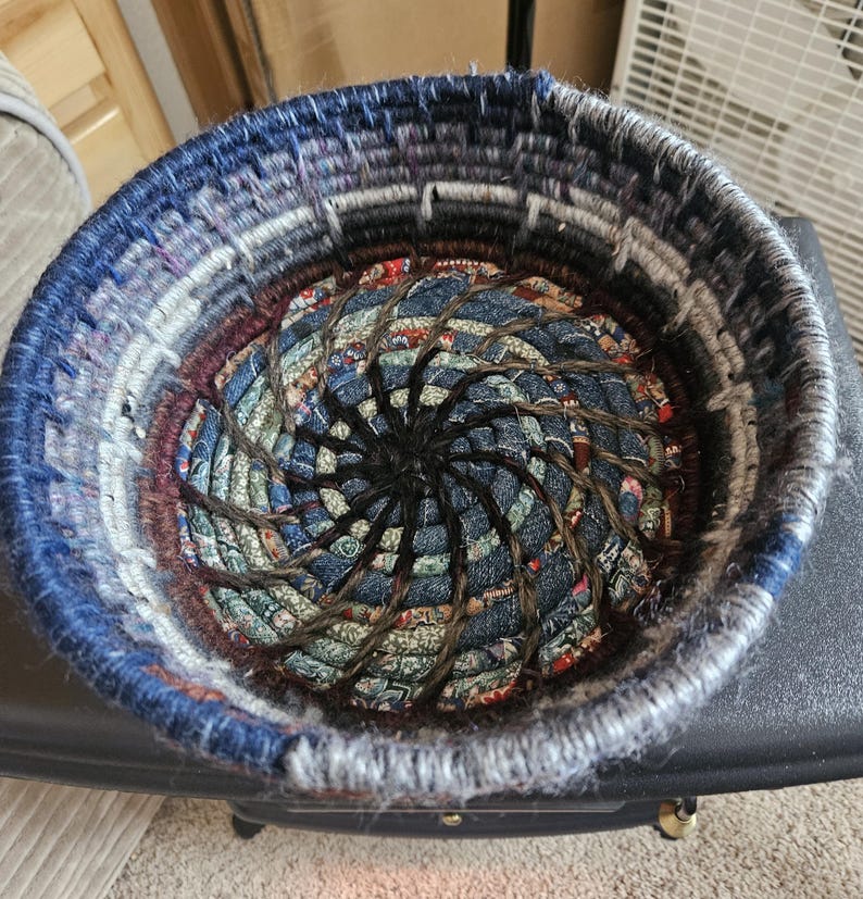 K&ouml;nnte beinhalten: Handgefertigter runder Korb mit Spiralmuster. Der Korb besteht aus Jeansstoff und anderen Stoffen in Blau-, Grau- und Burgundert&ouml;nen. Der Innenraum weist ein Spiralmuster mit dunkelbraunem Seil auf.