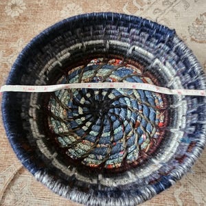 K&ouml;nnte beinhalten: Ein runder, gewebter Korb mit einem Durchmesser von etwa 18 cm. Der Korb besteht aus Denim- und Stoffstreifen in Blau-, Grau- und Braunt&ouml;nen. Das Zentrum hat ein Spiralmuster.