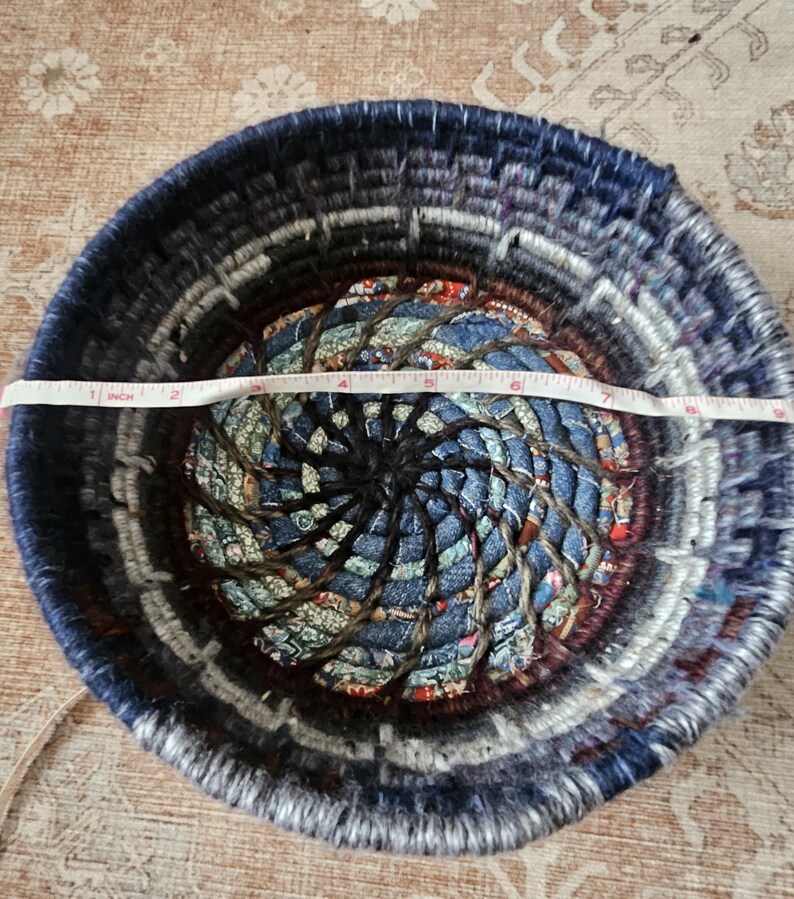 K&ouml;nnte beinhalten: Ein runder, gewebter Korb mit einem Durchmesser von etwa 18 cm. Der Korb besteht aus Denim- und Stoffstreifen in Blau-, Grau- und Braunt&ouml;nen. Das Zentrum hat ein Spiralmuster.