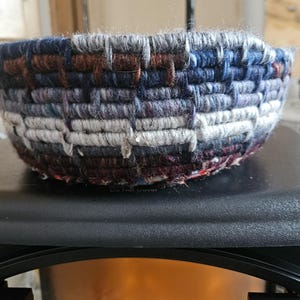 K&ouml;nnte beinhalten: Eine gewebte Schale mit strukturierter Oberfl&auml;che. Die Schale besteht aus Garn in den Farbt&ouml;nen Blau, Grau, Braun und Lila. Die Schale ist rund und hat eine weite &Ouml;ffnung.