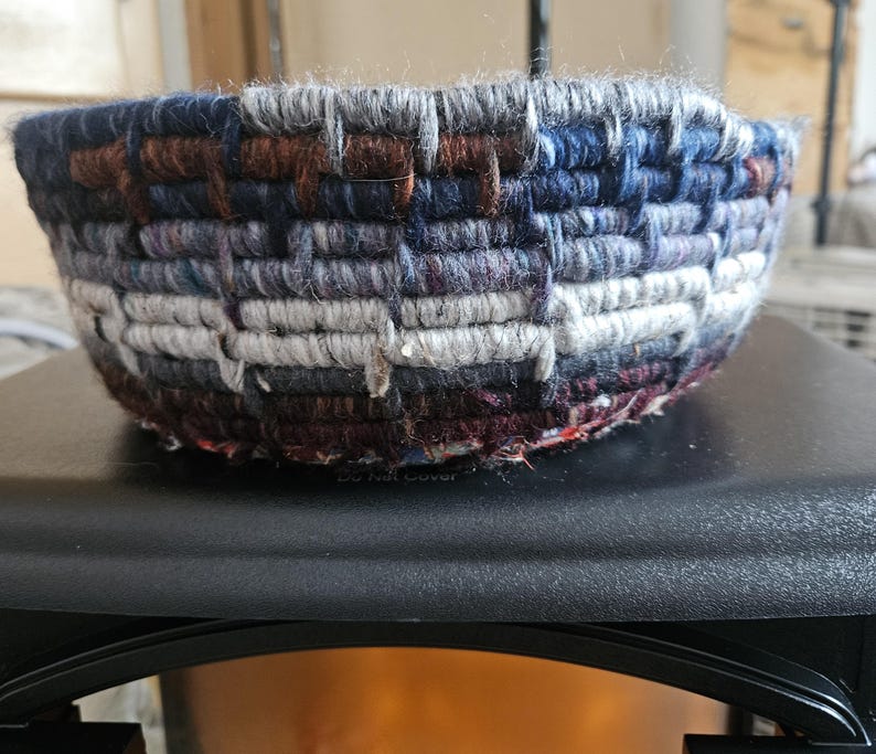 K&ouml;nnte beinhalten: Eine gewebte Schale mit strukturierter Oberfl&auml;che. Die Schale besteht aus Garn in den Farbt&ouml;nen Blau, Grau, Braun und Lila. Die Schale ist rund und hat eine weite &Ouml;ffnung.