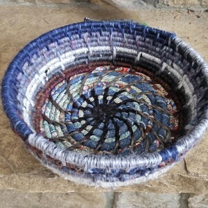 K&ouml;nnte beinhalten: Ein runder, gewebter Korb mit einem Spiralmuster in der Mitte. Der Korb hat eine Farbpalette aus Blau-, Grau- und Burgundert&ouml;nen. Der Rand ist tiefblau und der Innenraum hat eine gemusterte Stoffbasis.