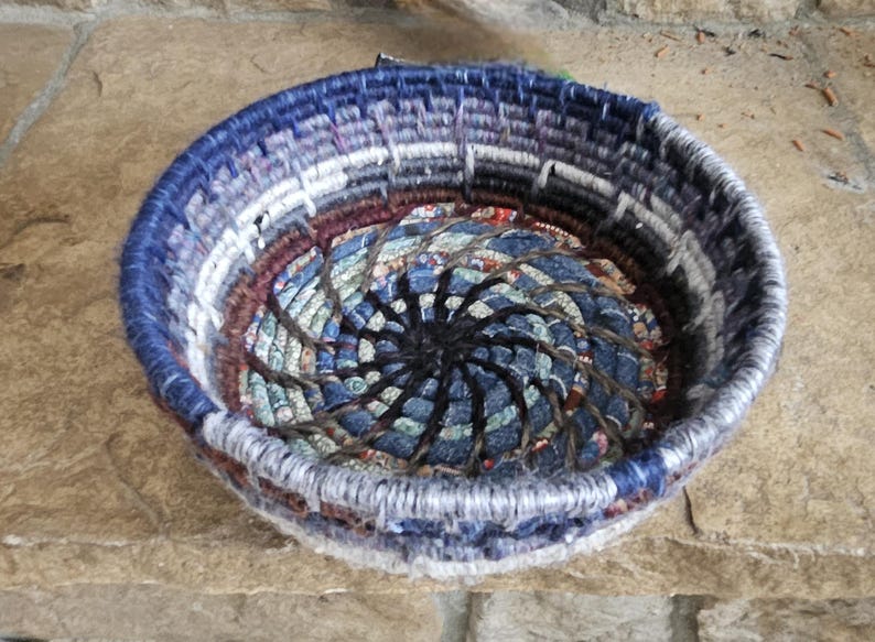 K&ouml;nnte beinhalten: Ein runder, gewebter Korb mit einem Spiralmuster in der Mitte. Der Korb hat eine Farbpalette aus Blau-, Grau- und Burgundert&ouml;nen. Der Rand ist tiefblau und der Innenraum hat eine gemusterte Stoffbasis.