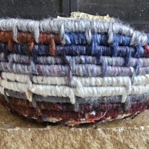 K&ouml;nnte beinhalten: Ein handgewebter Korb mit runder Form. Der Korb besteht aus dickem, strukturiertem Garn in den Farbt&ouml;nen Wei&szlig;, Grau, Blau, Braun und Lila. Das gewebte Design erzeugt ein gestreiftes Muster um die Au&szlig;enseite des Korbes. Der Korb steht auf einer Steinoberfl&auml;che.