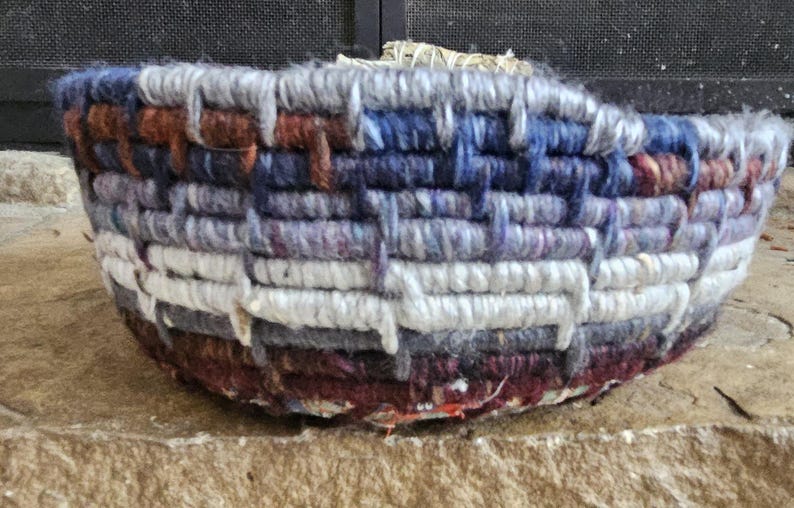 K&ouml;nnte beinhalten: Ein handgewebter Korb mit runder Form. Der Korb besteht aus dickem, strukturiertem Garn in den Farbt&ouml;nen Wei&szlig;, Grau, Blau, Braun und Lila. Das gewebte Design erzeugt ein gestreiftes Muster um die Au&szlig;enseite des Korbes. Der Korb steht auf einer Steinoberfl&auml;che.