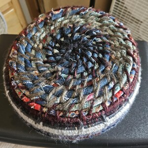 K&ouml;nnte beinhalten: Ein runder, handgefertigter Untersetzer aus geflochtenen Stoffresten. Der Untersetzer hat ein Spiraldesign mit Denim-, roten und gemusterten Stoffstreifen. Die Kanten sind mit einer dunkelburgunderfarbenen und wei&szlig;en Bord&uuml;re versehen. Durchmesser ca. 10 cm.